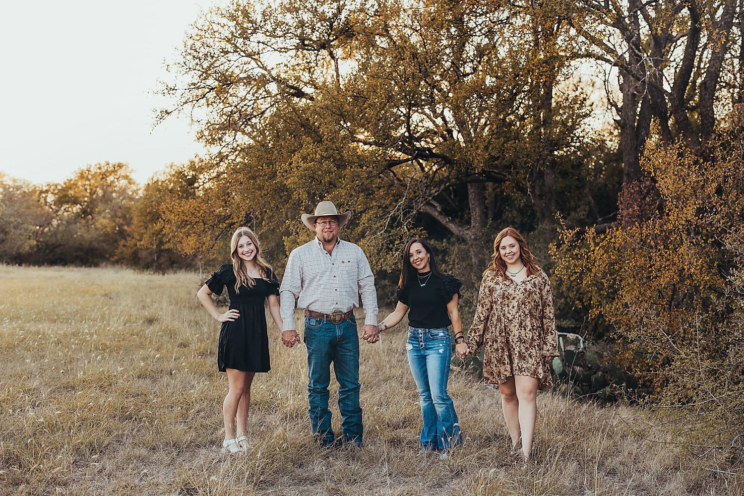 family photo with trees,grass in background. female in black dress, male longsleeve white shirt, jeans, boots and cowboy hat,female black shirt, jeans,boots, female brown floral dress and boots